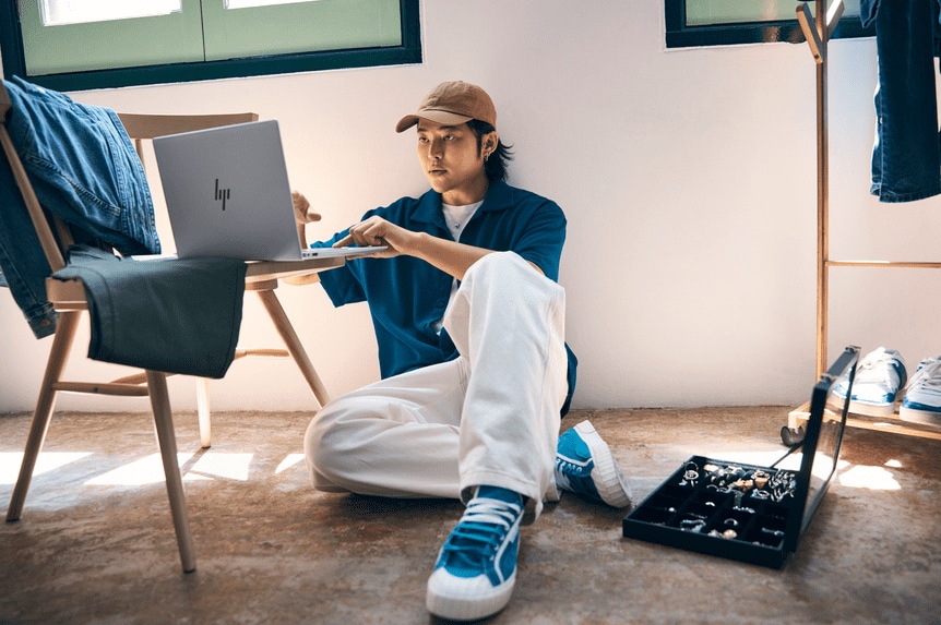 A young person wearing a brown cap, blue shirt, and white pants sits on the floor using an HP laptop.